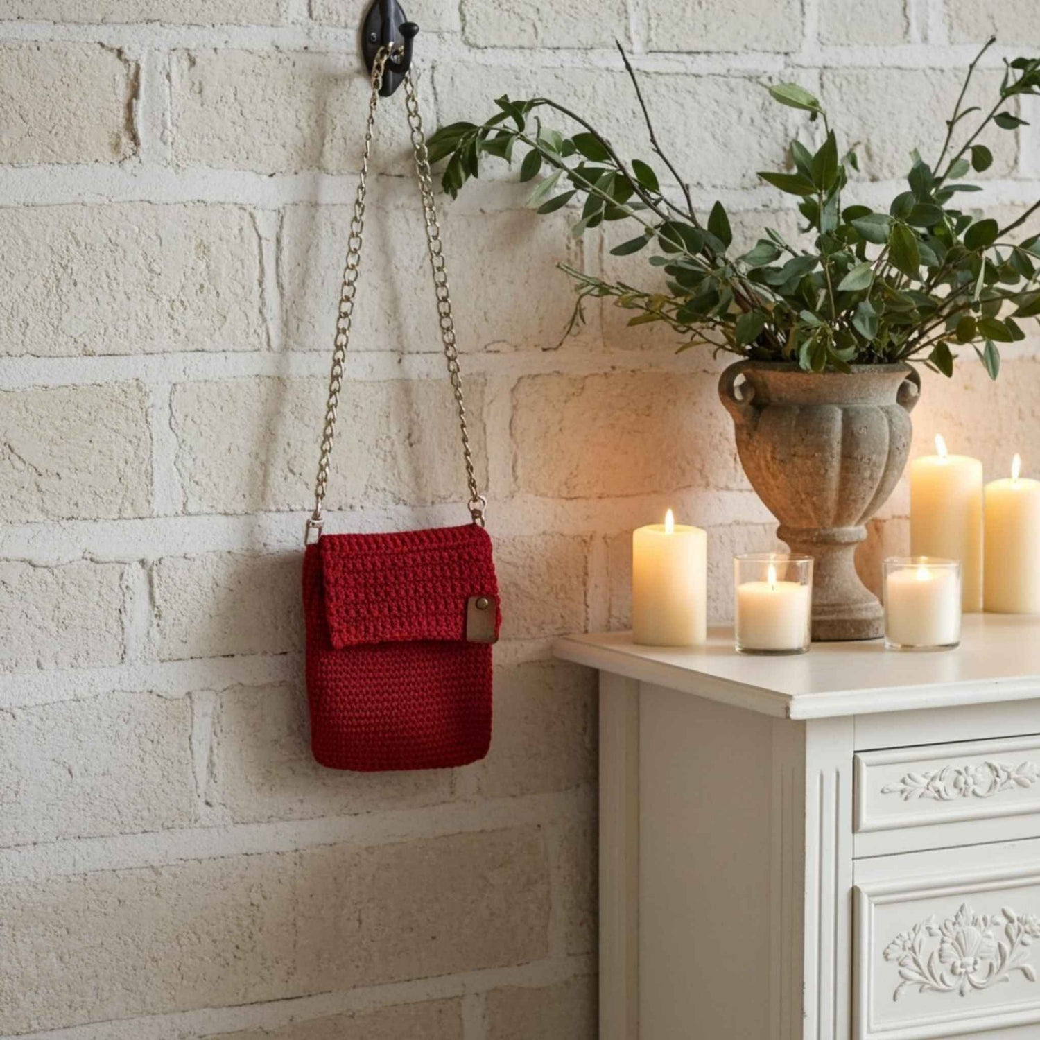 Red crocheted phone bag hanging on a brick wall next to a white cabinet with candles and a plant.