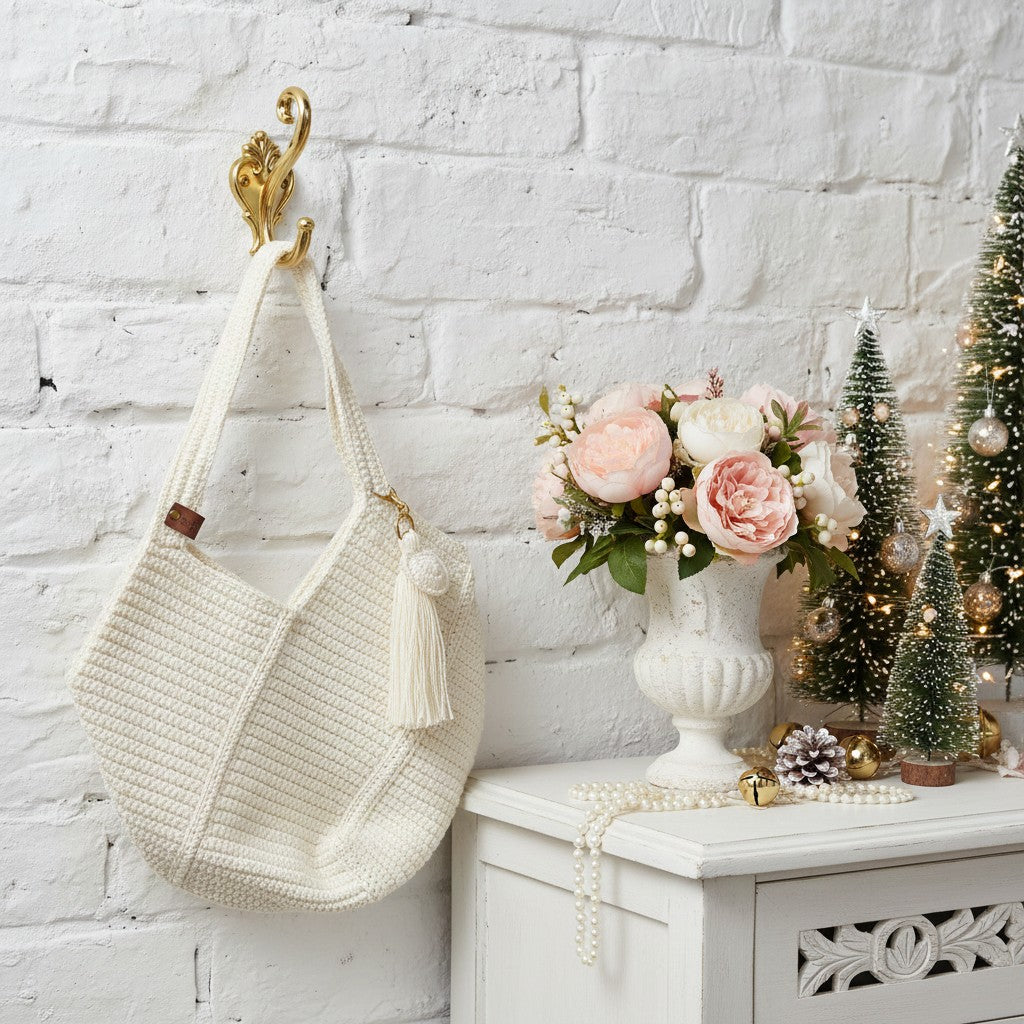 Crocheted bag with tassels hanging on a hook next to a vase of flowers and small Christmas trees against a white brick wall.