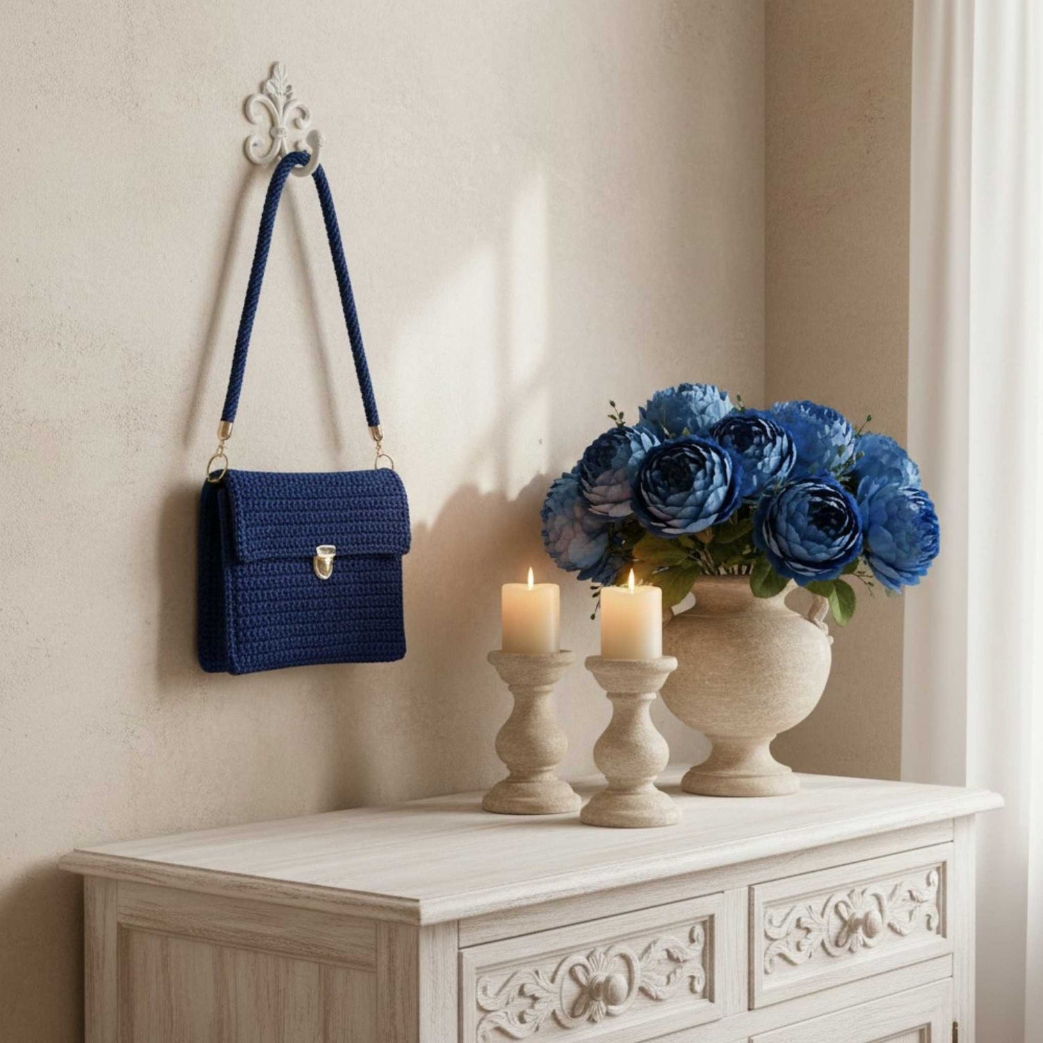 Blue handbag hanging on a wall hook above a decorative cabinet with candles and flowers.