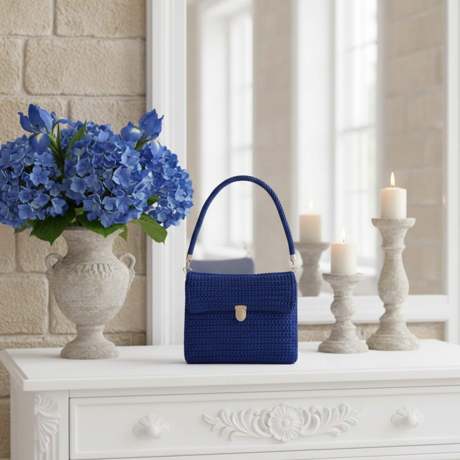 Blue handbag on a white surface with a vase of blue flowers and candles in the background.