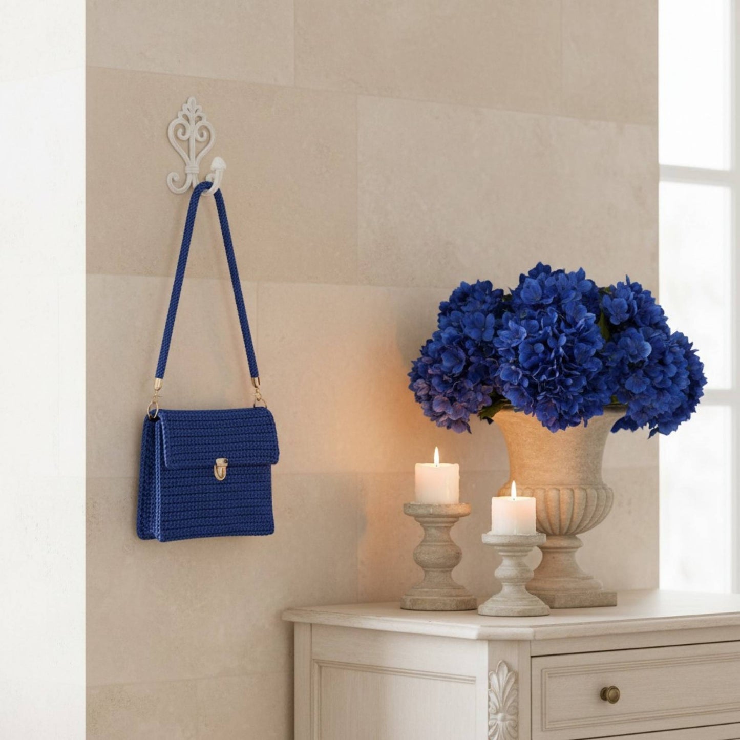 Blue handbag hanging on a wall hook next to a vase of blue flowers and candles on a cabinet.