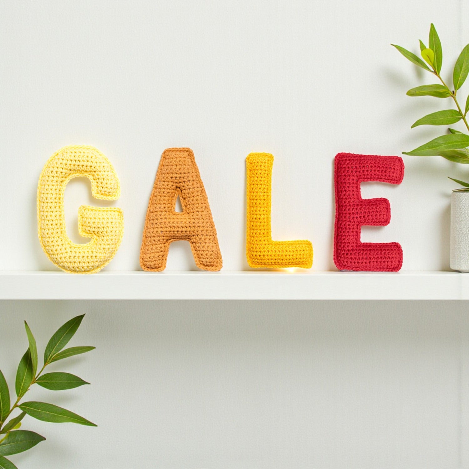 Crocheted letters spelling 'GALE' on a white shelf with green plants.
