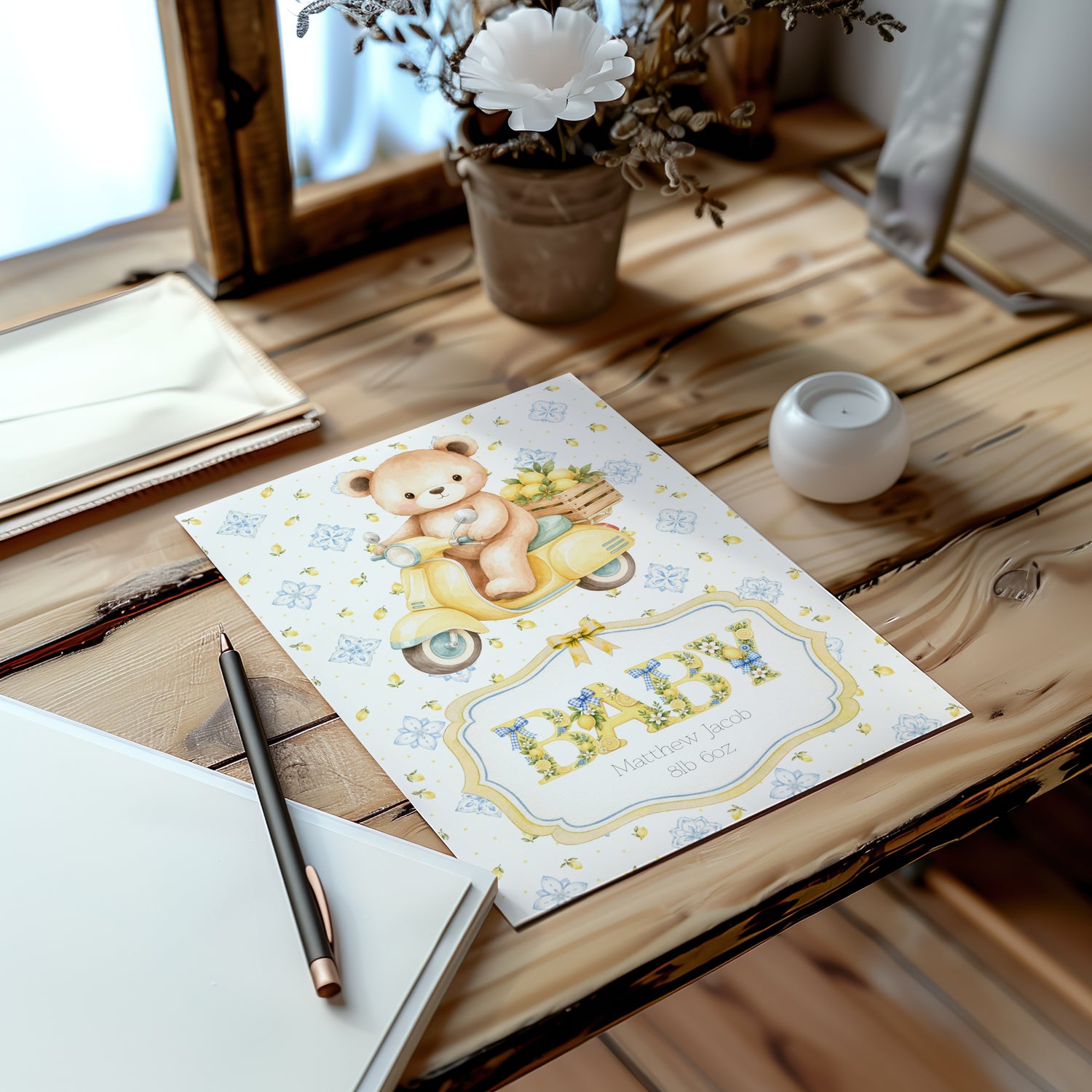 A baby card featuring a teddy bear on a yellow scooter and the word 'BABY' in decorative lettering, styled on a rustic wooden table with a pen, notebook, candle, and dried flowers.