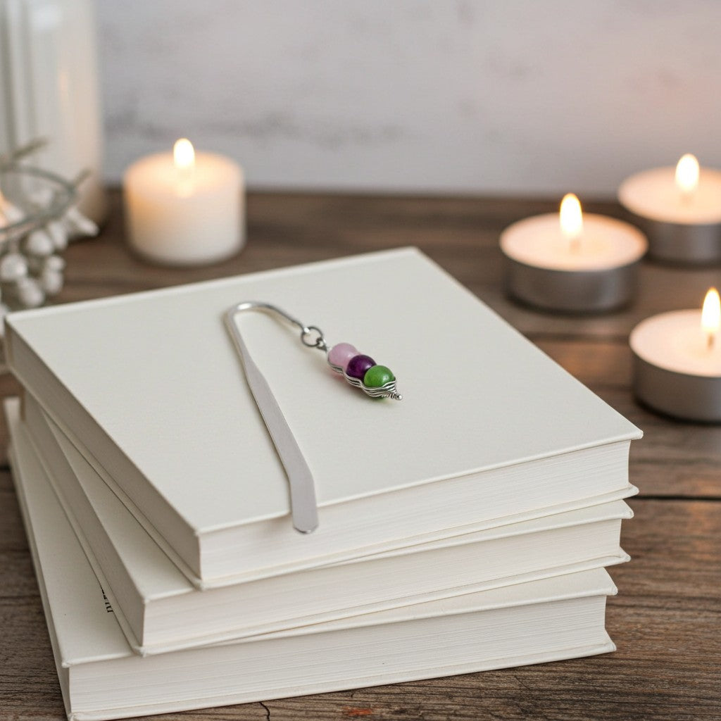 A silver metal bookmark with three small beads (purple, pink, and green) rests on a stack of white hardcover books on a wooden table, with lit candles blurred in the background.