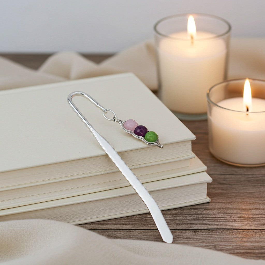 A silver metal bookmark with three small oval gemstones in pink, purple, and green, resting on a stack of cream-coloured books beside two lit candles on a wooden surface.
