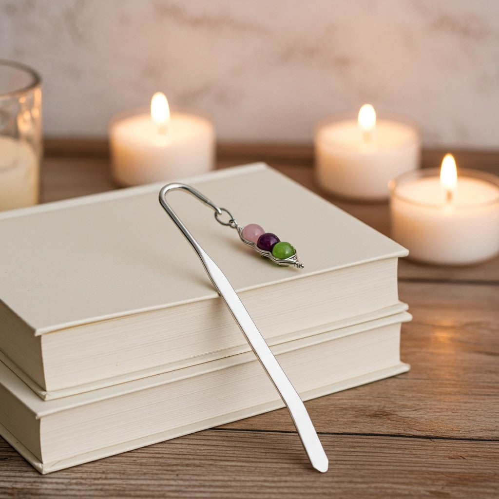 A silver metal bookmark with three small gemstone beads (pink, purple, and green) resting on a stack of cream-coloured books on a wooden table, with three lit candles blurred in the background.