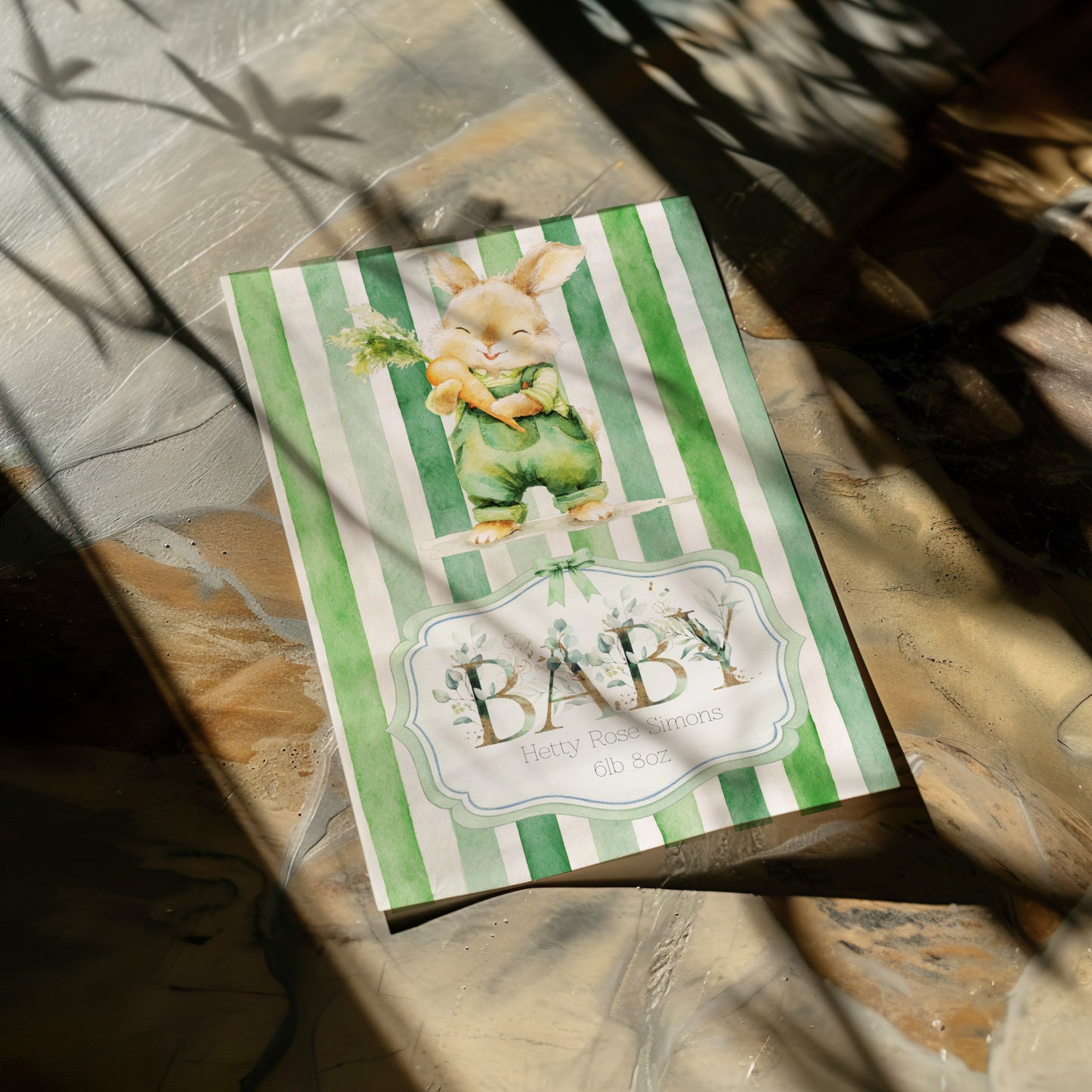 A baby card featuring a green and white striped background with a cute bunny illustration and the text 'BABY Baby shower' on a marble surface with dappled sunlight.
