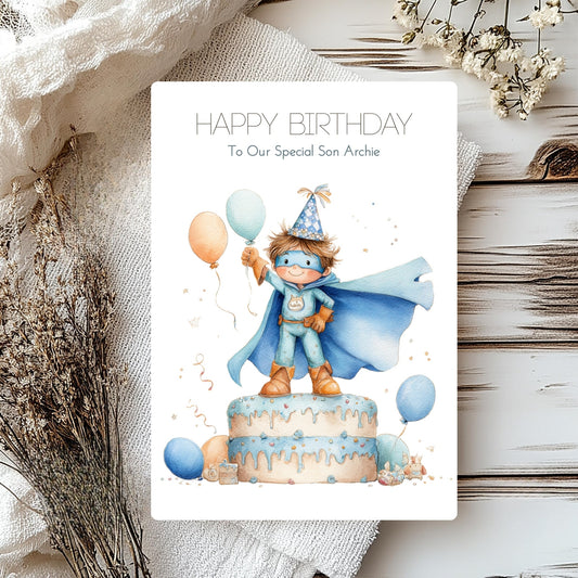 Personalised son card of a boy in a cape on top of a cake and balloons.