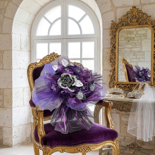 A purple and silver artificial flower bouquet with tulle wrapping and small white leaf accents, displayed on an ornate purple velvet and gold chair in a classical stone room with a mirror and console table.
