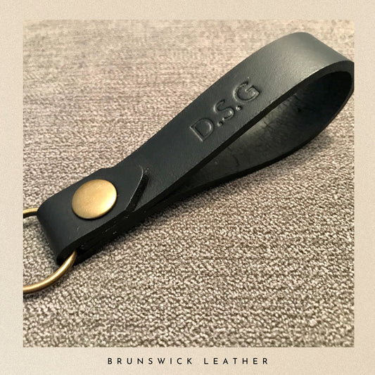 Black leather keychain with gold accent and 'D.S.G' engraving on a textured beige background.
