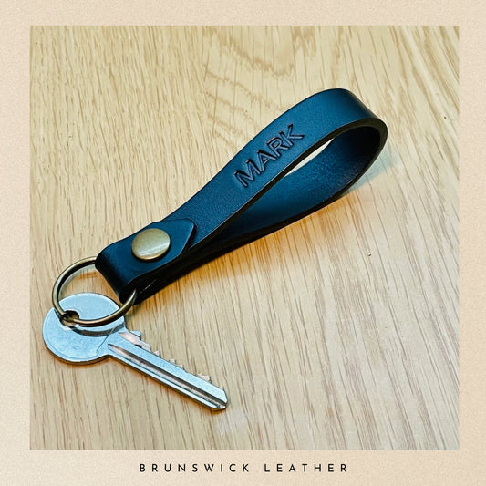 Black leather keychain with 'MARK' engraving on a wooden surface, branded 'Brunswick Leather'.