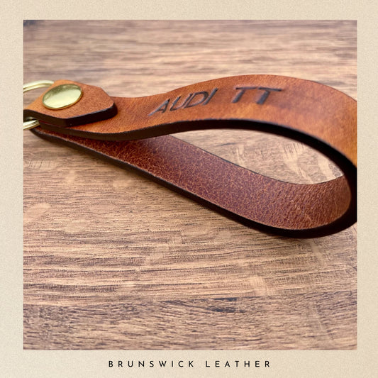 Brown leather keychain with 'Audi TT' engraving on a wooden surface, branded 'Brunswick Leather'.