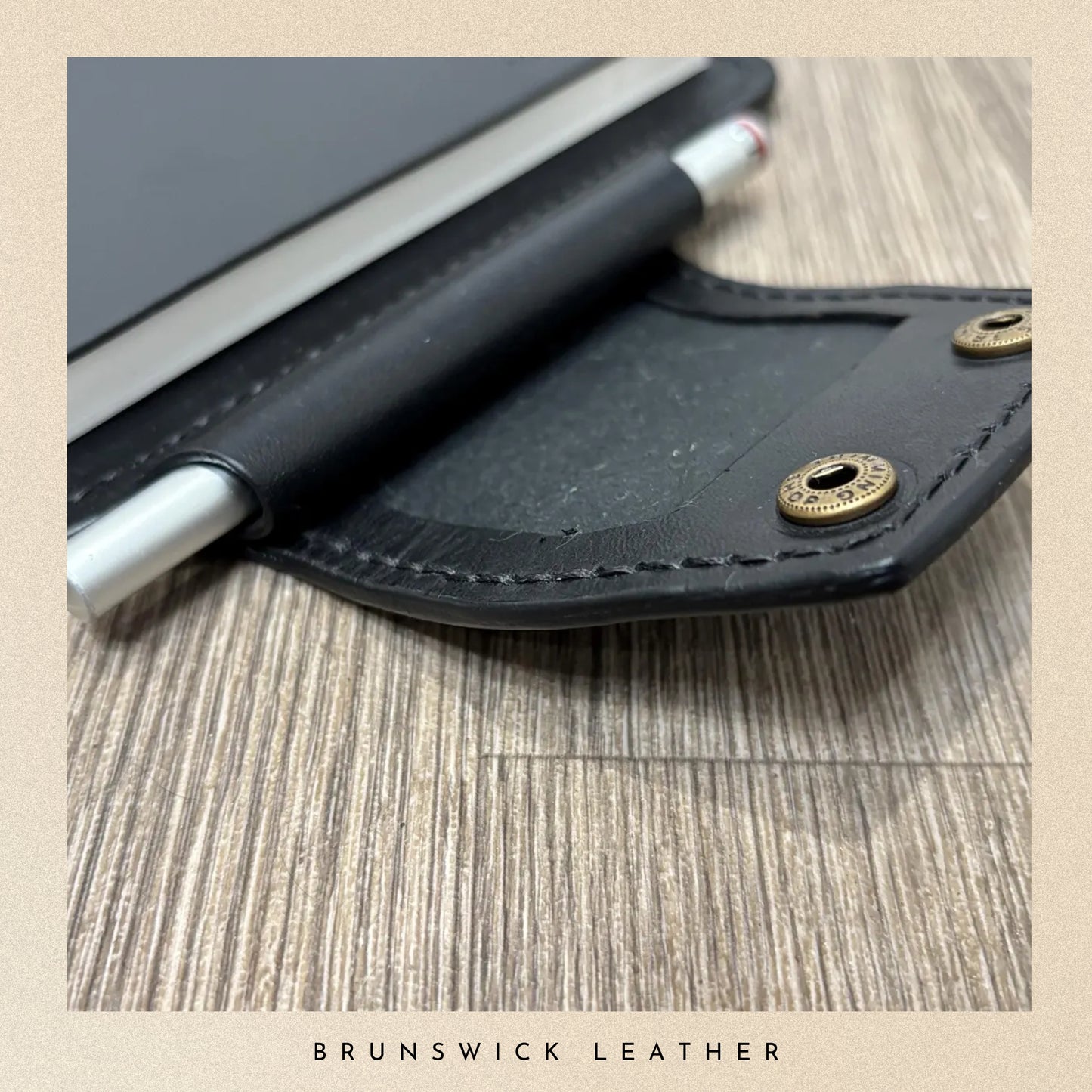 Black leather case with pen on a wooden surface, branded 'Brunswick Leather'.