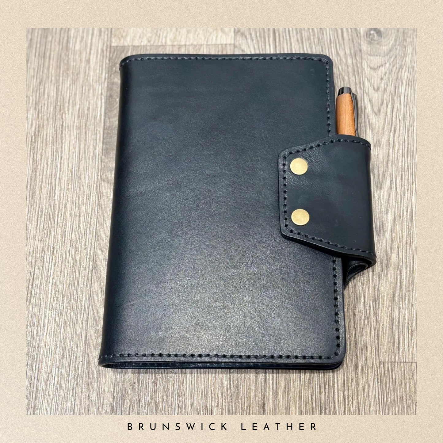 Black leather notebook with a pen holder on a wooden surface, branded 'Brunswick Leather'.