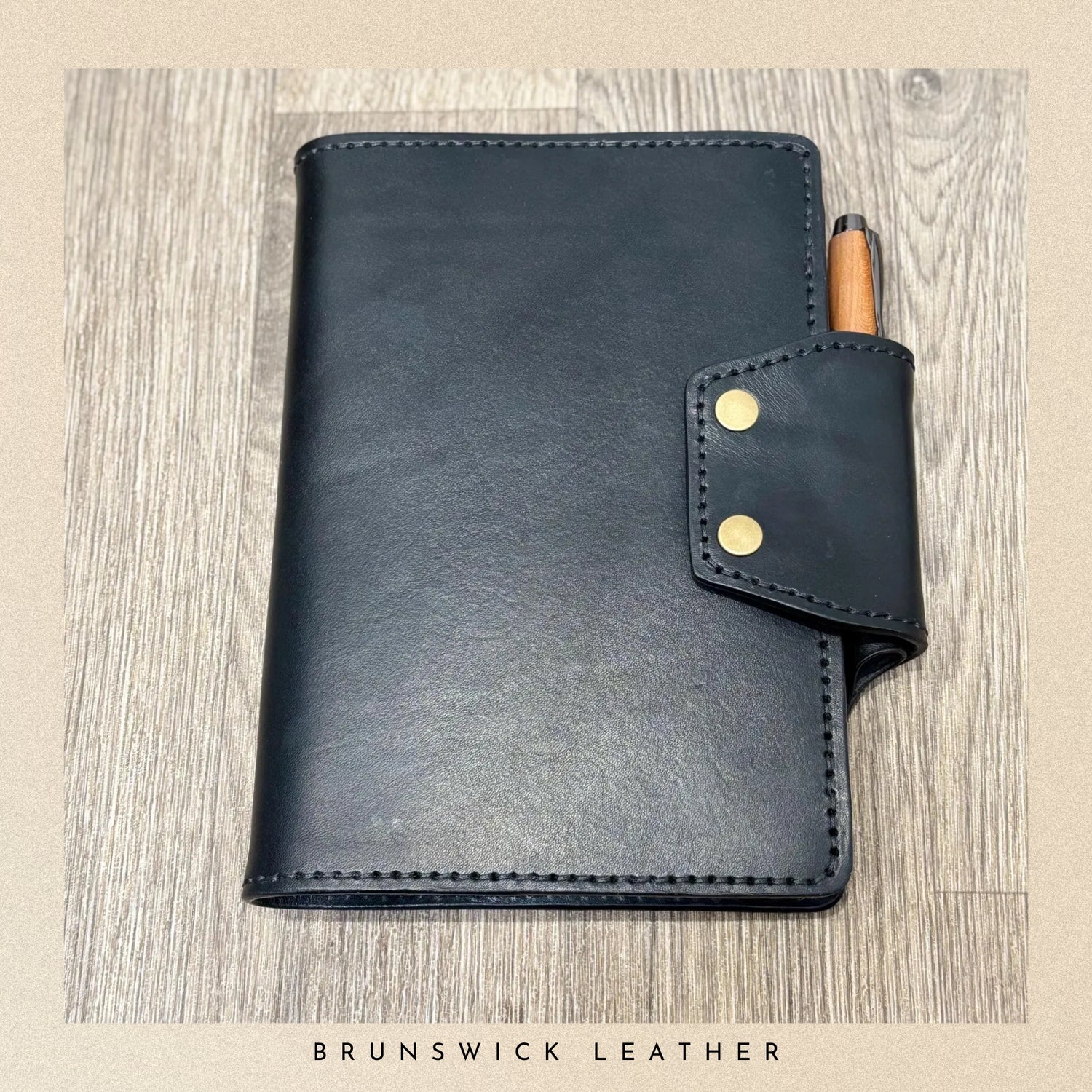 Black leather notebook with a pen holder on a wooden surface, branded 'Brunswick Leather'.