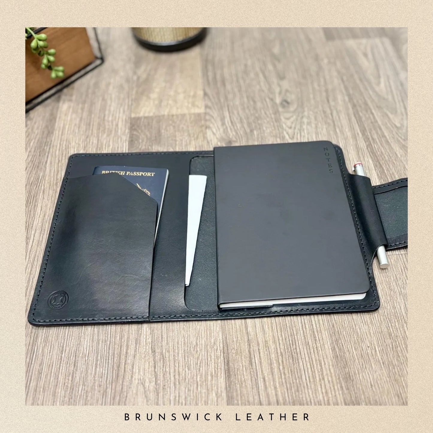 Black leather travel wallet with passport holder and pen on a wooden surface, branded 'Brunswick Leather'.