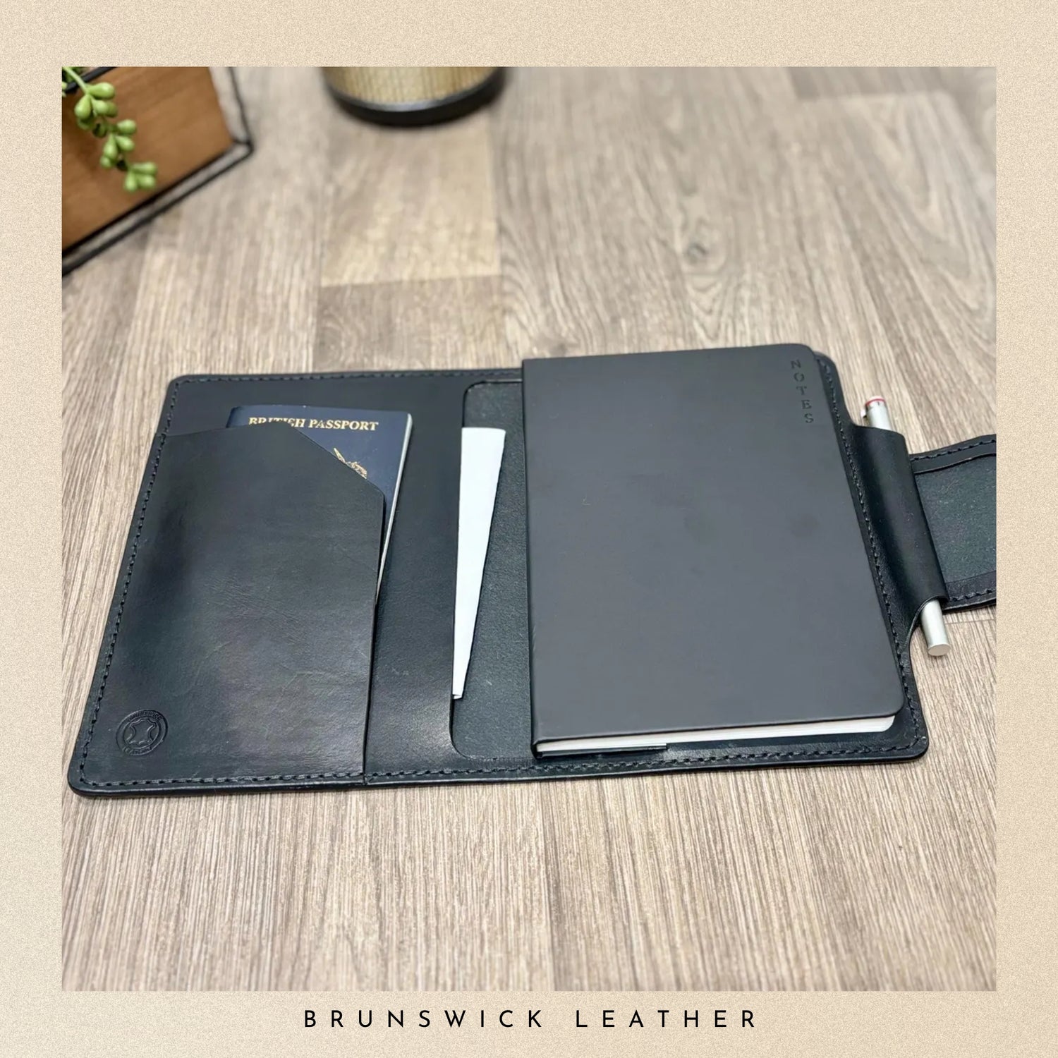 Black leather travel wallet with passport holder and pen on a wooden surface, branded 'Brunswick Leather'.