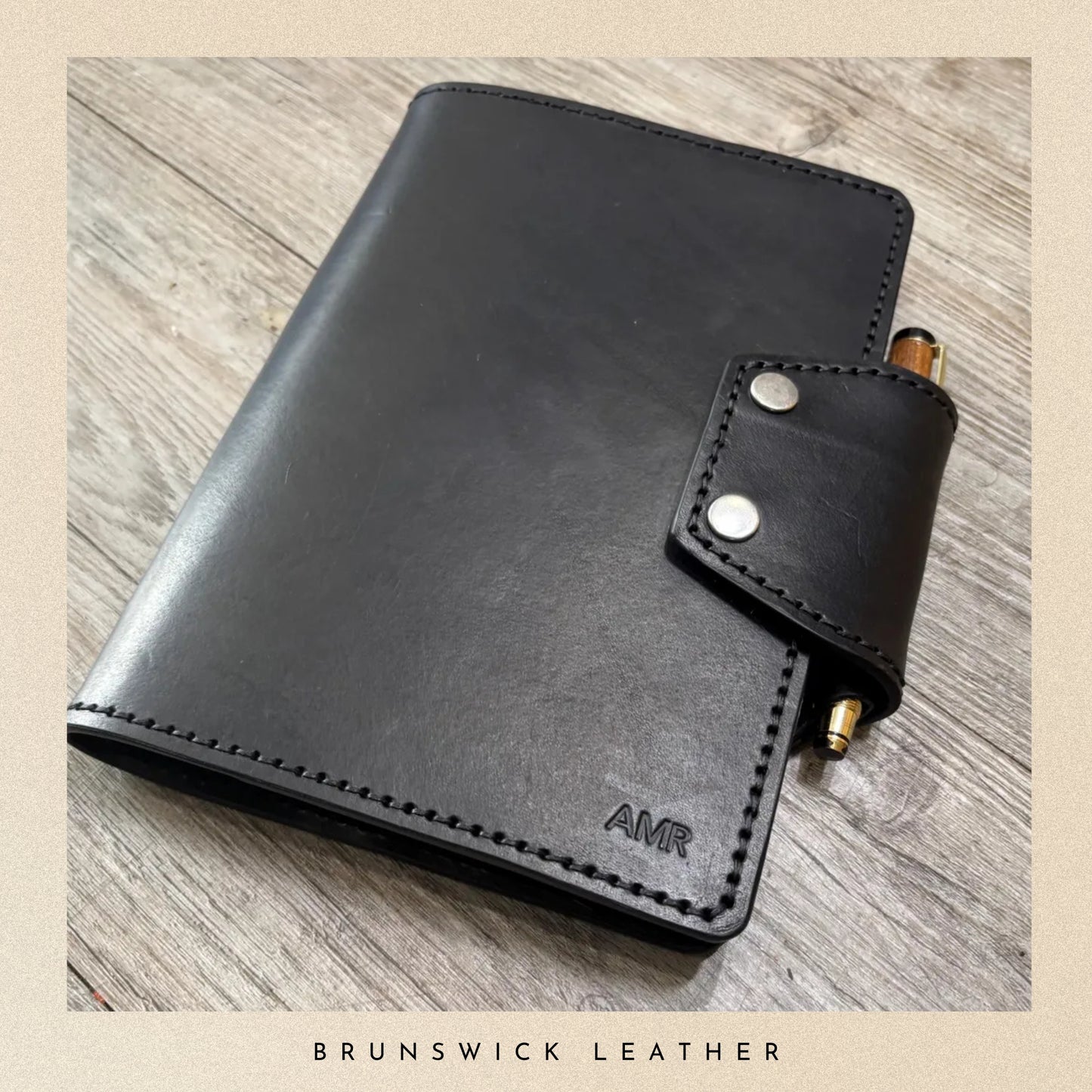 Black leather notebook with a pen holder on a wooden surface, branded 'AMR'.