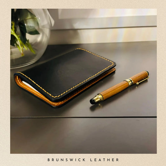 Black leather wallet and wooden pen on a reflective surface with 'Brunswick Leather' branding.