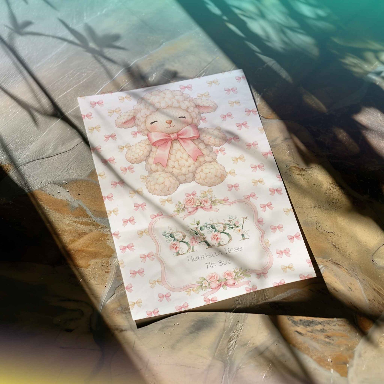 A decorative card featuring a cute cream-coloured lamb with a pink bow, surrounded by small pink bows and floral motifs, placed on a stone surface with dappled sunlight and shadows.