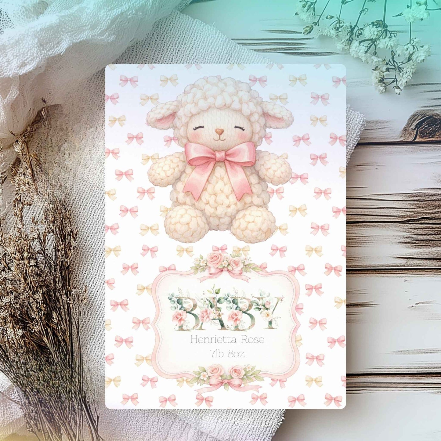 A baby shower card featuring a cute cream-colored woolly lamb with a pink bow, surrounded by a pattern of small bows. The bottom of the card has a decorative frame with the word 'BABY' in floral letters and the personalized name 'Henrietta Rose' with baby weight