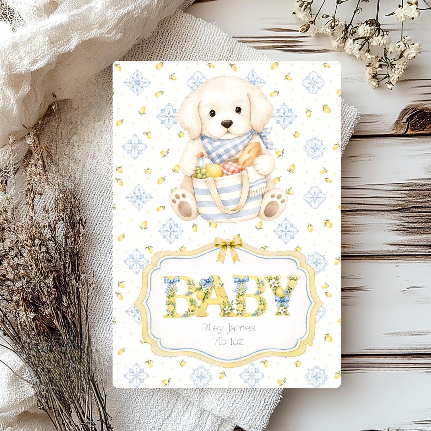 A baby shower card featuring a cute puppy illustration holding a basket of treats, with the word 'BABY' in floral lettering and the names 'Riley James' and '7th 1oz' printed below. The card is styled on a rustic white wood surface with a white textured cloth, dried flowers, and a small wooden slice.