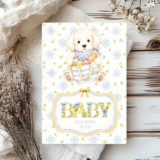 A baby shower card featuring a cute puppy illustration holding a basket of treats, with the word 'BABY' in floral lettering and the names 'Riley James' and '7th 1oz' printed below. The card is styled on a rustic white wood surface with a white textured cloth, dried flowers, and a small wooden slice.