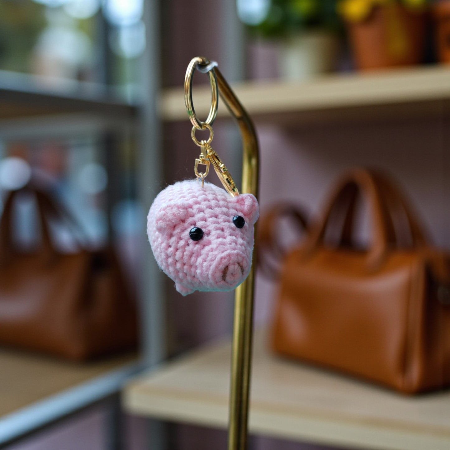 Pink crochet pig keychain on a gold hook with blurred handbags in the background