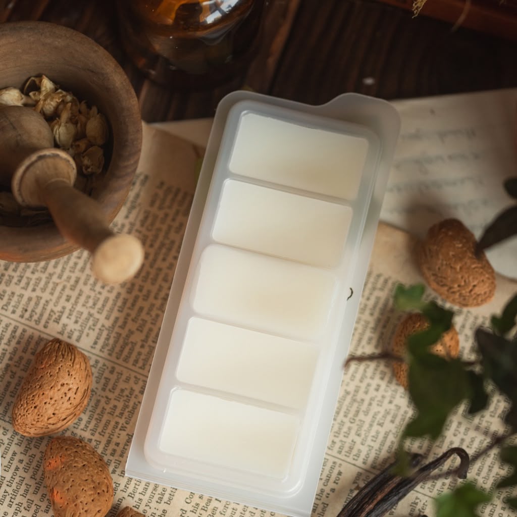 A translucent silicone mold with five rectangular cavities, filled with white wax melts, styled on a vintage book page with botanical and craft props.