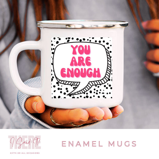 A white ceramic enamel mug with black text and design, held in a person's hand. The mug has a speech bubble graphic with the phrase 'YOU ARE ENOUGH' and dots around the edge.