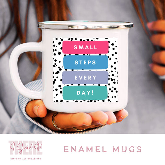 A white ceramic enamel mug with a handle, featuring a pattern of colourful text that reads 'SMALL STEPS EVERY DAY!' , held in a person's hand.