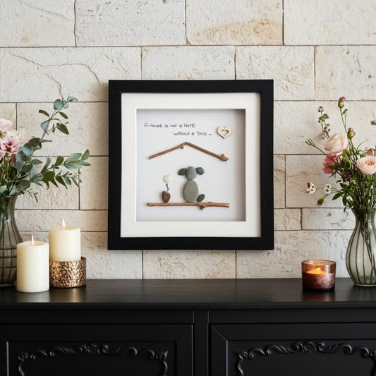 A framed pet memorial wall art piece displayed on a black console table against a light stone wall. The black-framed shadow box features a white mat, a small grey pebble dog under a twig house, a heart-shaped stone, and the printed message 'A house is not a home without a dog.
