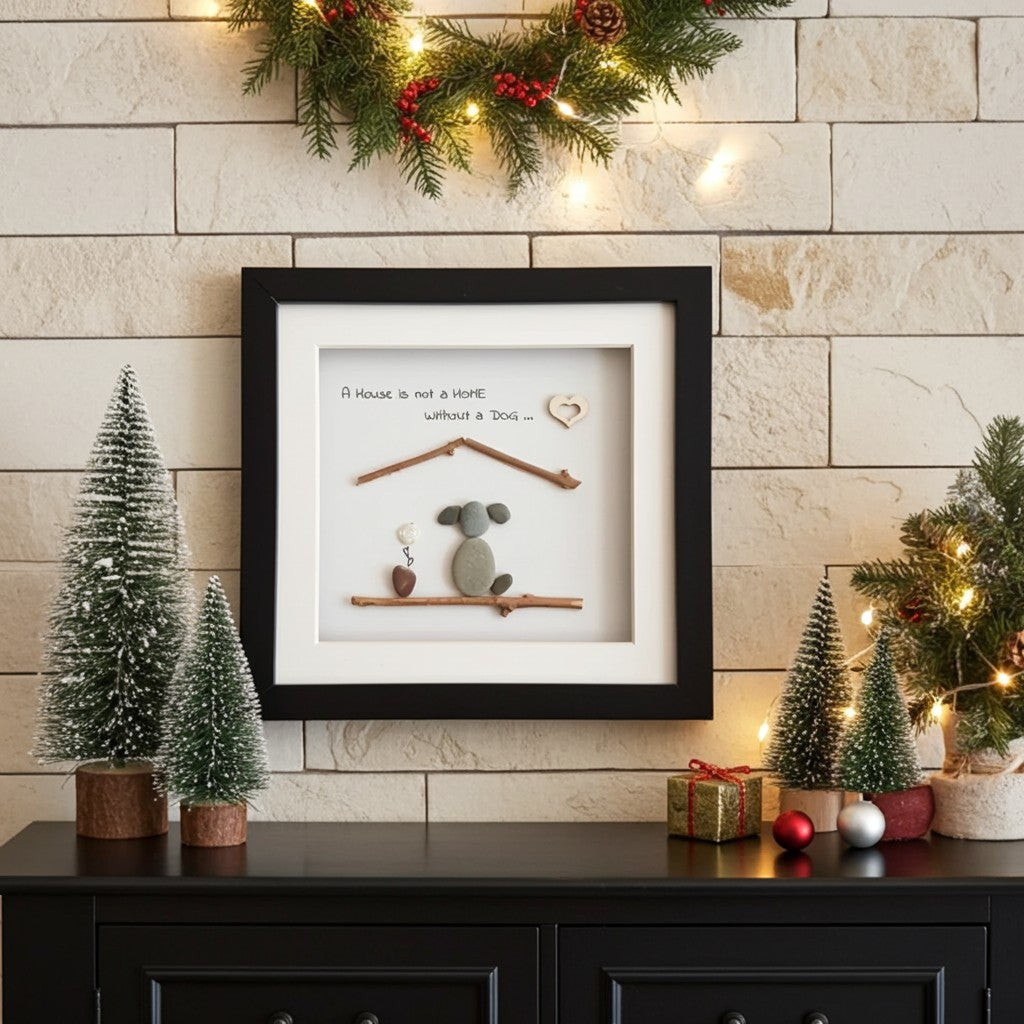 A framed custom pet pebble wall art piece displayed on a black console table against a light stone brick wall, decorated with miniature Christmas trees, a garland, and warm string lights. The frame contains a white background with a black shadow box effect, featuring a small grey pebble dog under a twig roof, a small white dog figurine, and the text 'A Heart is not a HOME without a Dog.