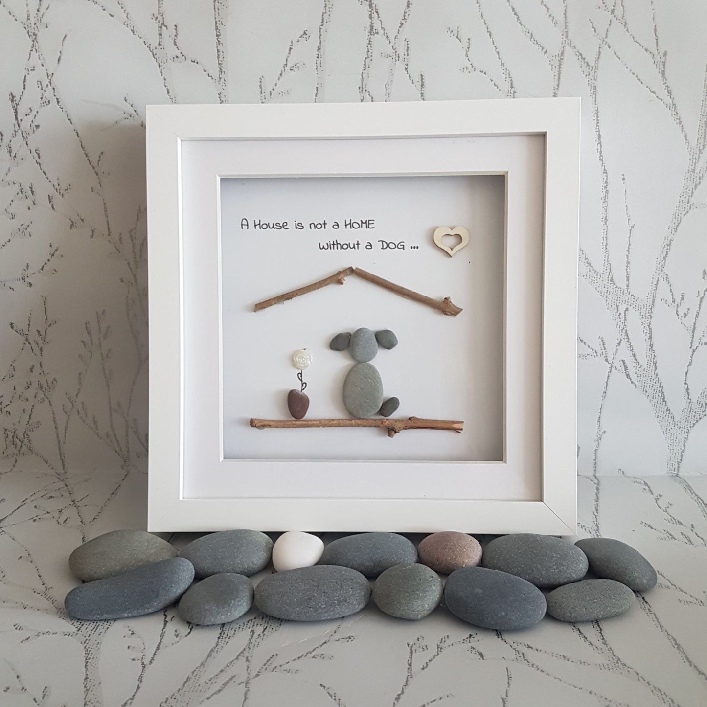 A pebble art picture frame containing a picture of a dog, a house, and some decorative pebbles in front of the frame. The frame has a white mat and a clear glass cover.