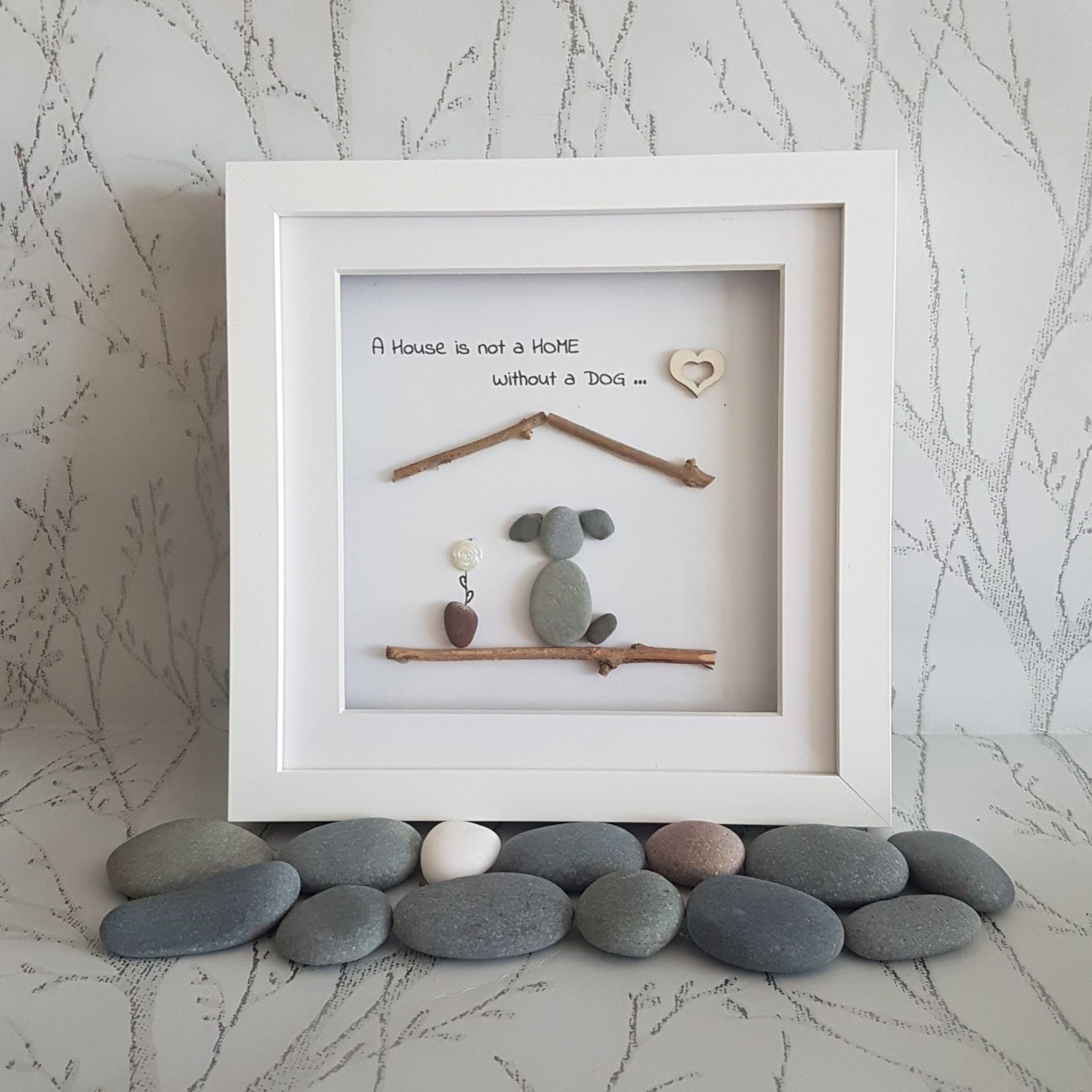 A pebble art picture frame containing a picture of a dog, a house, and some decorative pebbles in front of the frame. The frame has a white mat and a clear glass cover.