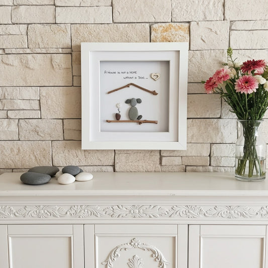 A framed handmade wall art piece displayed on a white console table against a light beige stone wall. The white square frame contains a small handcrafted scene with natural stones, twigs, a tiny heart, and the handwritten quote 'A house is not a home without a dog.