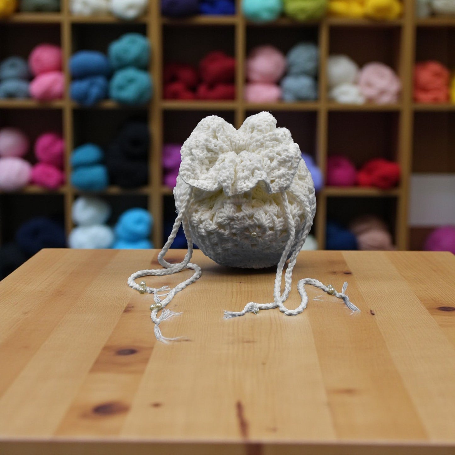 White drawstring crochet bag on a wooden table with colorful yarn balls in the background