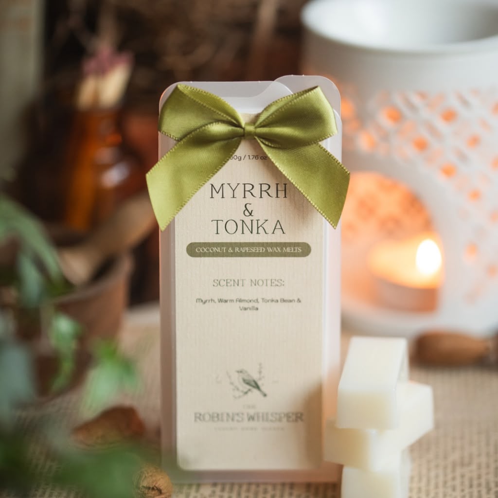 A boxed scented wax melt package labeled 'MYRRH & TONKA' by Robin Whisper, tied with an olive green ribbon, standing on a burlap surface with candles and warm-toned decor blurred in the background.