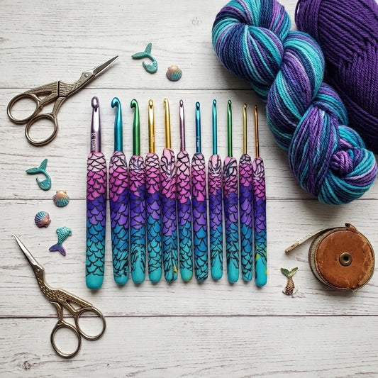 A set of nine crochet hooks with colourful mermaid-scale handles in purple, pink, and turquoise, arranged on a white wooden surface with yarn skeins, scissors, shells, and a spool as props.