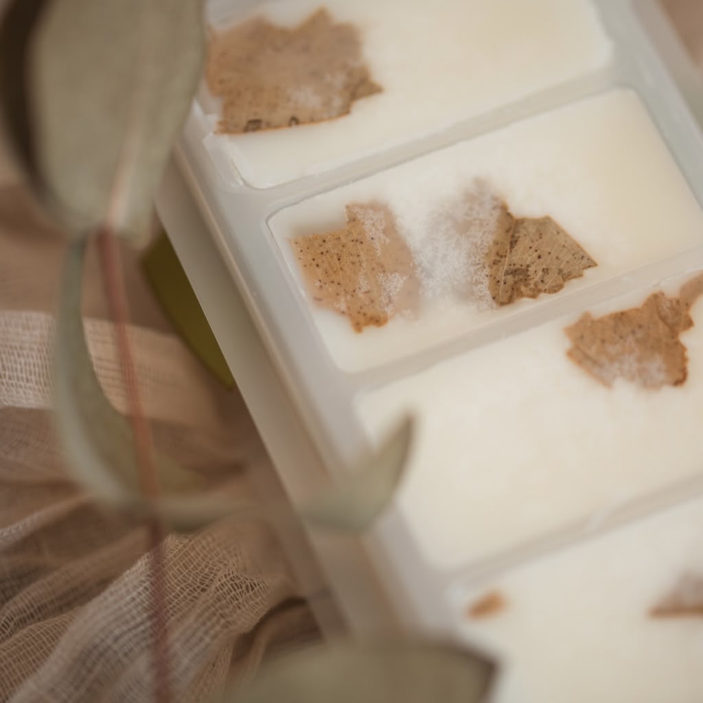 A close-up of a white tray containing several rectangular wax melts, each with a small dried botanical piece embedded on top. The scene is softly lit and styled with blurred fabric and leaves in the foreground.