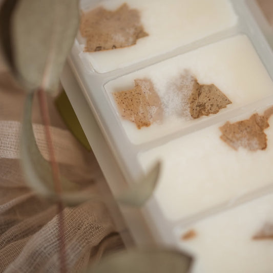 A close-up of a white tray containing several rectangular wax melts, each with a small dried botanical piece embedded on top. The scene is softly lit and styled with blurred fabric and leaves in the foreground.
