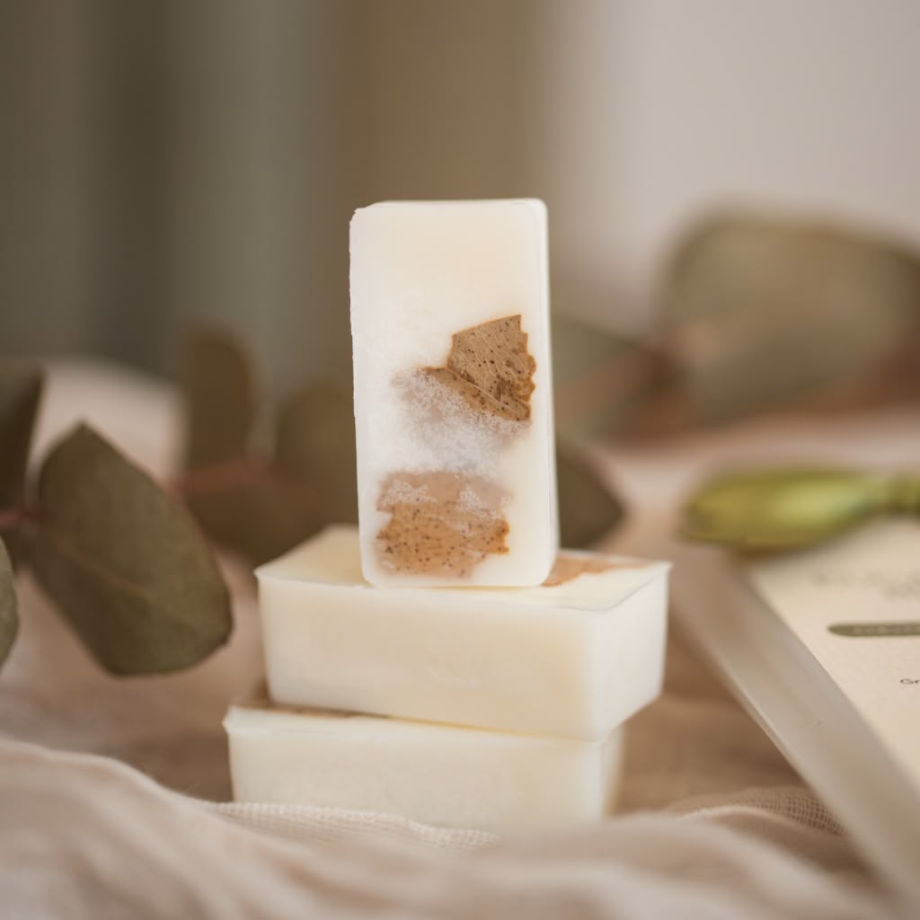 Three stacked rectangular bars of wax melts, with the top bar standing upright to show embedded brown and white botanical pieces, styled with dried leaves and a book in a soft neutral setting.