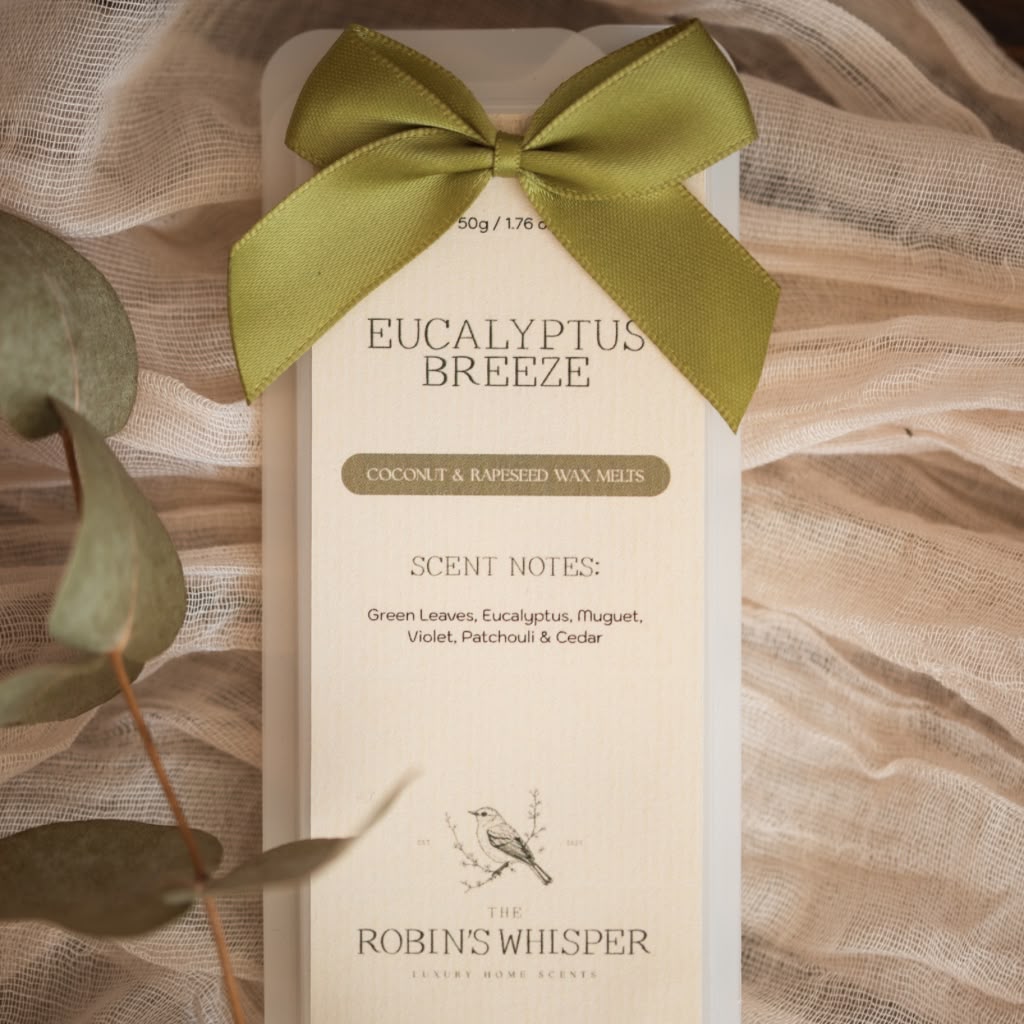 A boxed wax melt package labeled 'Eucalyptus Breeze' by Robin's Whisper, tied with an olive green satin bow, photographed on draped beige fabric with eucalyptus leaves around it.