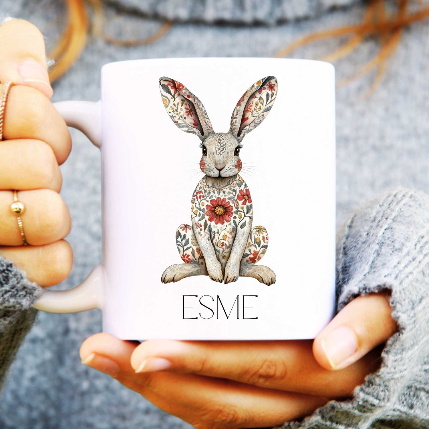 White mug with a decorative hare design and 'ESME' text held by hands.