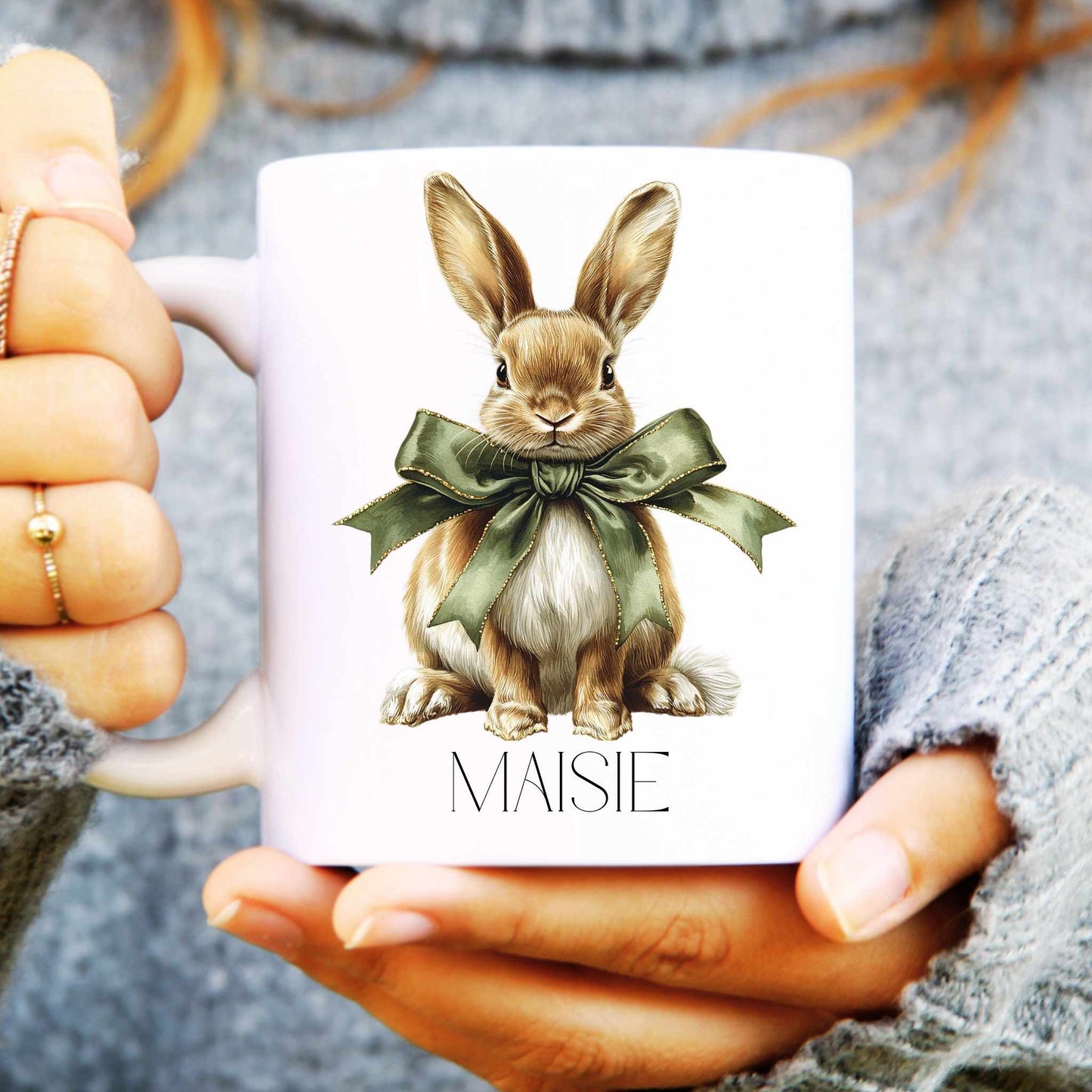 White mug with a hare illustration and 'MAISIE' text held by hands.