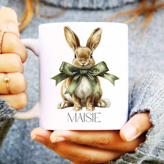 White mug with a hare  illustration and 'MAISIE' text held by hands.