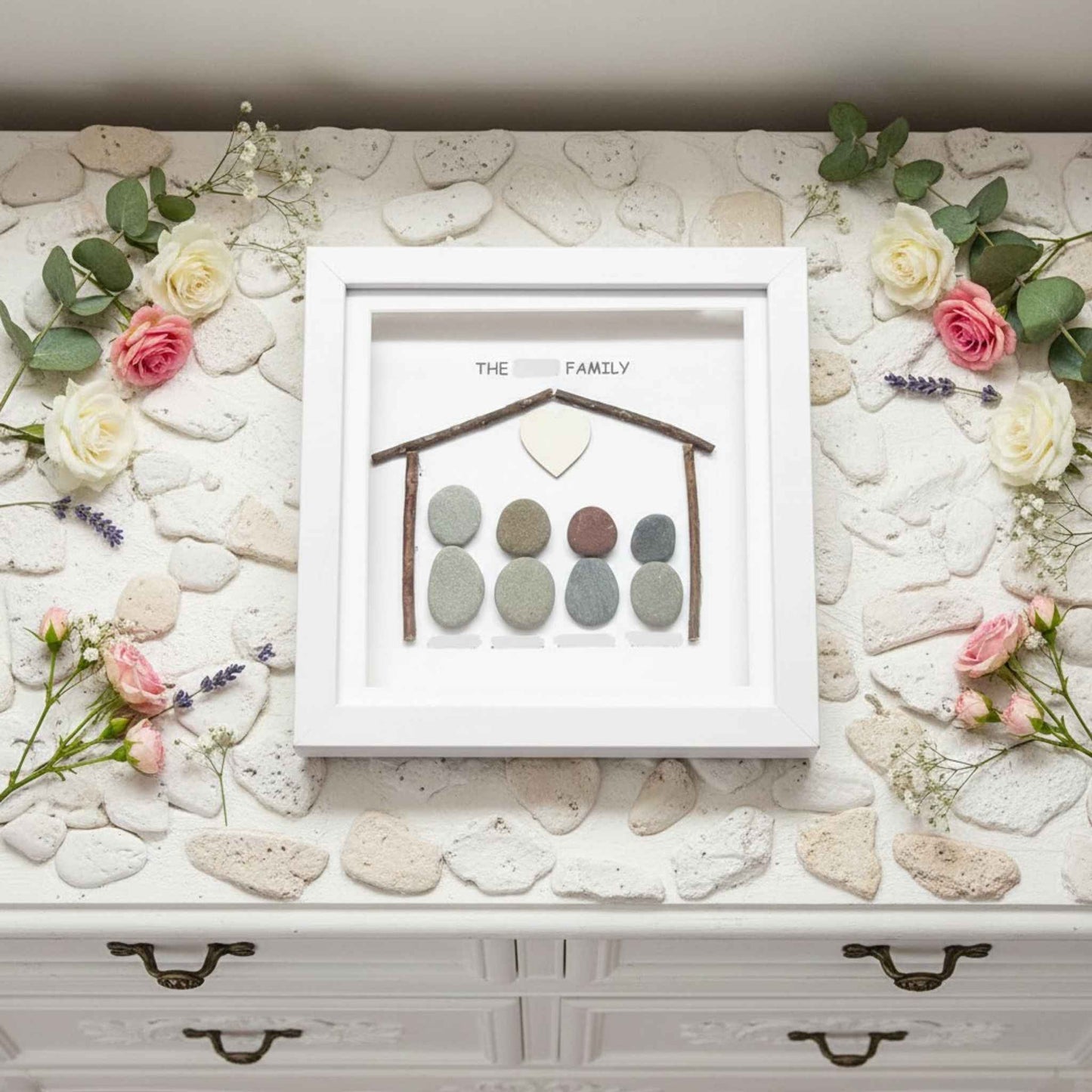 Decorative display with pebble art in a white frame on a stone surface with flowers.