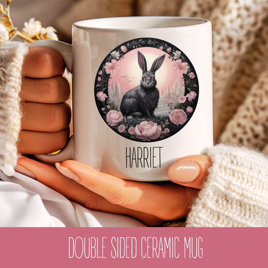 A ceramic coffee mug with a double-sided design featuring a fantasy hare (rabbit) with pink flowers and a cityscape in the background, and the name 'Harriet' at the bottom.