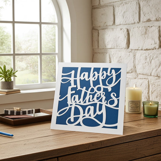 A square greeting card with white cursive script reading 'Happy Father's Day' on a blue background, displayed on a wooden table in a bright room with a window, brick wall, candle, and small plant.