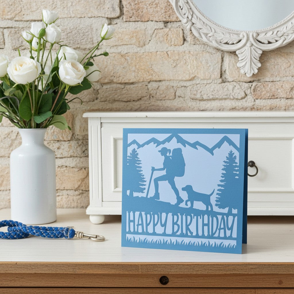 A blue and light blue layered greeting card with a silhouette of a hiker and dog in front of mountains, reading 'HAPPY BIRTHDAY', displayed on a wooden surface next to a white vase of white roses.